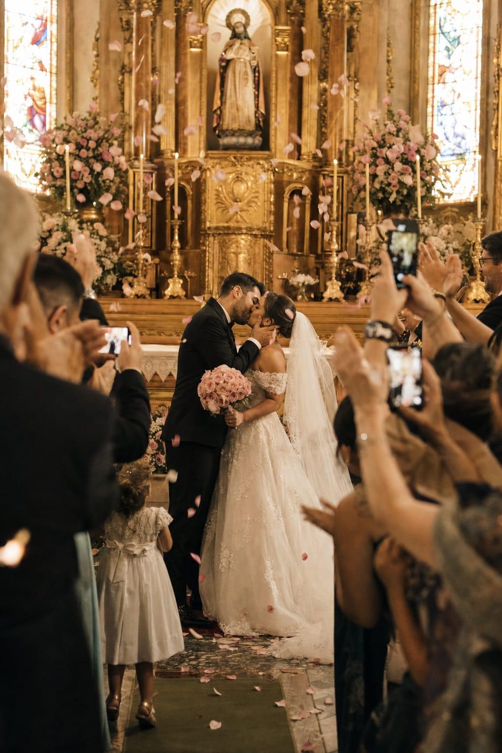 Beijo dos noivos na igreja, com chuva de pétalas e convidados fotografando